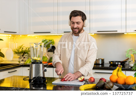 Middle-aged man making apple smoothie in kitchen listening to music on headphones for healthy diet Middle-aged man making apple smoothie in kitchen listening to music on headphones for healthy diet 137725737