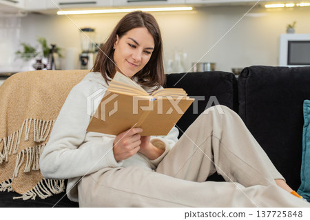Young woman enjoys leisure reading paper book smiles in comfort turns pages with bookmark calm ease Young woman enjoys leisure reading paper book smiles in comfort turns pages with bookmark calm ease 137725848