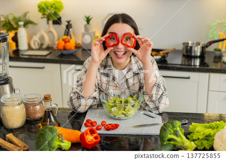 Playful young woman in apron making pepper glasses while preparing keto vegetable salad at home 137725855