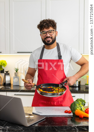 Arabian young adult man tossing hot vegetables in frying pan while watching laptop recipe in kitchen Arabian young adult man tossing hot vegetables in frying pan while watching laptop recipe in kitchen 137725864
