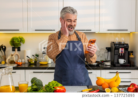 Middle-aged man celebrating big smartphone victory in home kitchen after online success notification Middle-aged man celebrating big smartphone victory in home kitchen after online success notification 137725893
