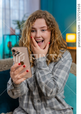 Young woman on sofa scrolling phone exclaiming wow discovering exciting discount or prize at home 137725916