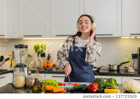 Young woman cutting cherry tomatoes listening to music preparing fresh vegetarian salad at home meal Young woman cutting cherry tomatoes listening to music preparing fresh vegetarian salad at home meal 137725920