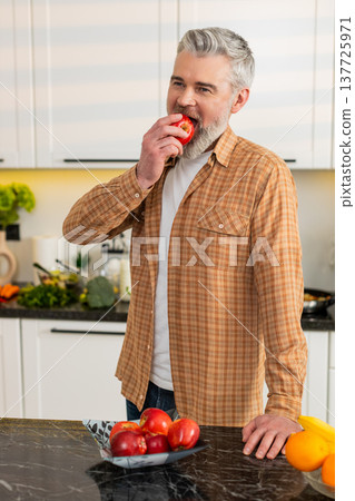 Middle-aged man bites fresh apple in kitchen for healthy diet snack smiling satisfied home kitchen 137725971