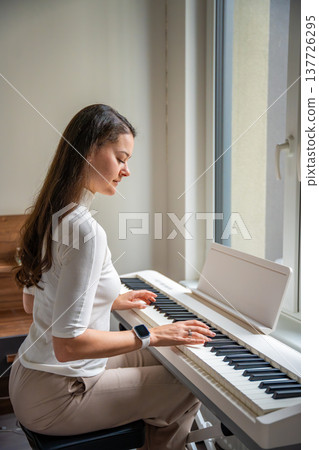 Young woman playing on white piano synthesizer at home. Concept of home lesson of music: learning music enhances motor skills, hearing, and intelligence, fostering creativity 137726295