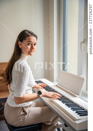 Young woman playing on white piano synthesizer at home. Concept of home lesson of music: learning music enhances motor skills, hearing, and intelligence, fostering creativity 137726297