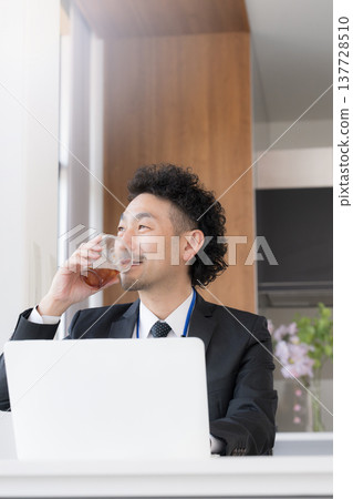 A bearded businessman, smiling and relaxed, is working at his desk, drinking coffee and using his laptop during a break. 137728510