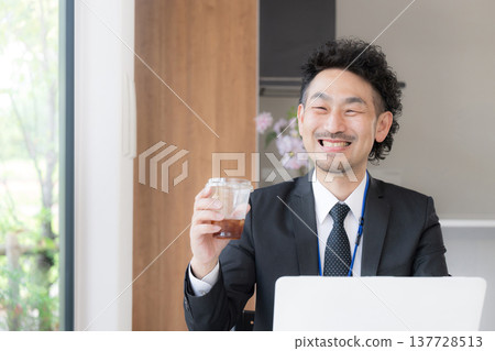 A bearded businessman, smiling and relaxed, is working at his desk, drinking coffee and using his laptop during a break. A bearded businessman, smiling and relaxed, is working at his desk, drinking coffee and using his laptop during a break. 137728513