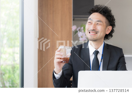 A bearded businessman, smiling and relaxed, is working at his desk, drinking coffee and using his laptop during a break. A bearded businessman, smiling and relaxed, is working at his desk, drinking coffee and using his laptop during a break. 137728514