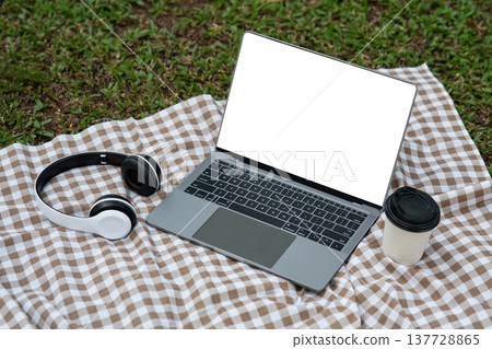 Modern workspace with laptop, headphones, and coffee cup on picnic blanket in green outdoor setting, showcasing lifestyle and productivity in nature Modern workspace with laptop, headphones, and coffee cup on picnic blanket in green outdoor setting, showcasing lifestyle and productivity in nature 137728865
