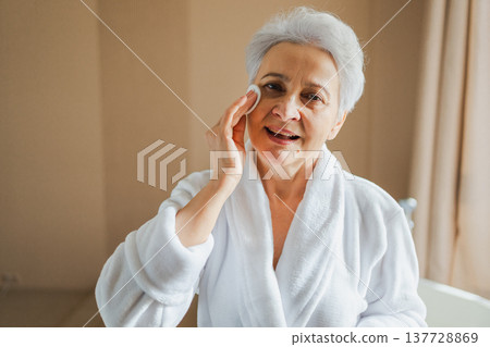 Mature adult old woman doing daily beauty routine removing makeup with cotton pad. Senior woman with lotion sponge washing face gets facial treatment. Skin care aging beauty skincare cleansing Mature adult old woman doing daily beauty routine removing makeup with cotton pad. Senior woman with lotion sponge washing face gets facial treatment. Skin care aging beauty skincare cleansing 137728869