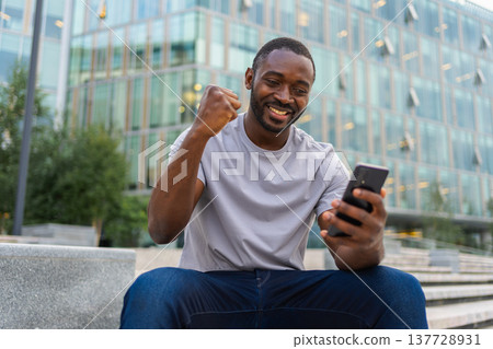 Happy African American man euphoric winner with smartphone on street in city. Person guy looking at cell phone reading great news getting good result winning online bid feeling amazed. Winning gesture 137728931
