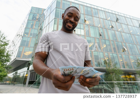 African American man holding money dollar bills on urban street in city. Businessman guy counting paper currency money outdoors near office bank building. Man holding cash. Finance economy banking 137728937