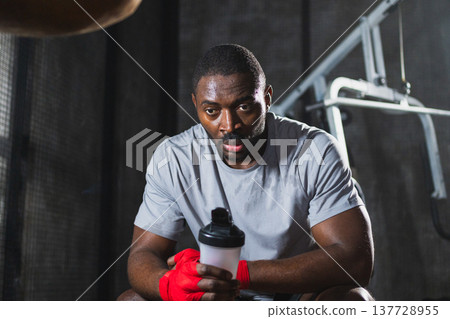 Boxer in gym. Tired African man fighter with red boxing wraps resting after boxing training session with boxing bag in gym. Strong sweaty athlete man after box sparring. Training day in gym 137728955