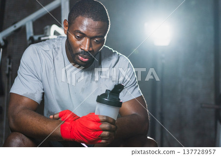 Boxer in gym. Tired African man fighter with red boxing wraps resting after boxing training session with boxing bag in gym. Strong sweaty athlete man after box sparring. Training day in gym Boxer in gym. Tired African man fighter with red boxing wraps resting after boxing training session with boxing bag in gym. Strong sweaty athlete man after box sparring. Training day in gym 137728957