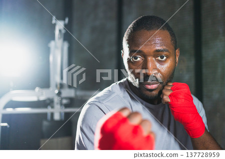 Workout in gym. African man fighter ready for fight punching with boxing wraps protective bandages to camera. Strong man training punches looking concentrated straight preparing for boxing sparring Workout in gym. African man fighter ready for fight punching with boxing wraps protective bandages to camera. Strong man training punches looking concentrated straight preparing for boxing sparring 137728959