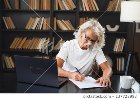 Middle aged senior woman using laptop computer writing notes at workplace. Focused mature old businesswoman work at office making written records doing online work. Mature boss professional worker Middle aged senior woman using laptop computer writing notes at workplace. Focused mature old businesswoman work at office making written records doing online work. Mature boss professional worker 137728988