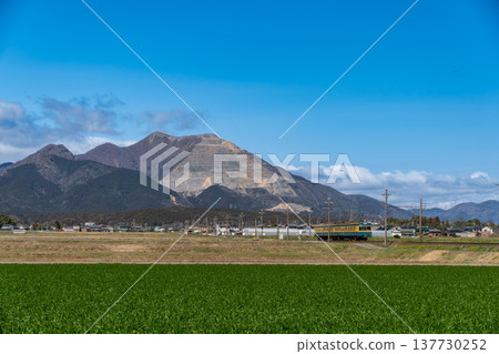 A green train on the Sangi Railway Sangi Line runs through a clear blue sky with Mount Fujiwara in the background. A green train on the Sangi Railway Sangi Line runs through a clear blue sky with Mount Fujiwara in the background. 137730252