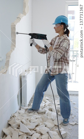 Woman construction worker demolishing a wall with a hammer drill, wearing protective gear, generating rubble pile during renovation project, vertical view Woman construction worker demolishing a wall with a hammer drill, wearing protective gear, generating rubble pile during renovation project, vertical view 137731367