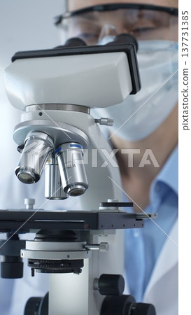 Scientist wearing lab coat and protective glasses using a modern microscope in a laboratory setting, conducting research and experiments in a scientific environment. Health care and science concept 137731385