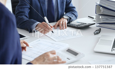 Two accountants examining financial documents using a calculator, taking notes, and discussing results, surrounded by documents and technology in a busy office environment. Audit and taxes in business 137731387