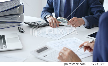 Male financial analysts with female colleague examining documents, using magnifying glass and calculator at workplace in office. Audit and taxes in business Male financial analysts with female colleague examining documents, using magnifying glass and calculator at workplace in office. Audit and taxes in business 137731398