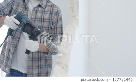 Male construction worker demolishing white wall with rotary hammer drill is wearing grey checkered shirt and protective white gloves, generating dust during renovation project, closeup view Male construction worker demolishing white wall with rotary hammer drill is wearing grey checkered shirt and protective white gloves, generating dust during renovation project, closeup view 137731445