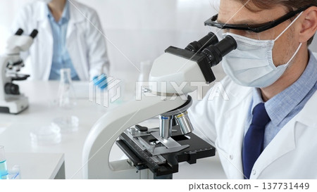 Masked scientist analyzing sample under microscope, colleague researching nearby in sterile medical laboratory setting. Medicine, healthcare and science concept 137731449