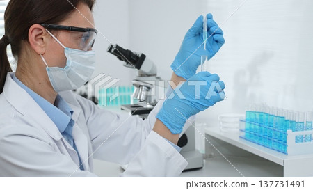 Female scientist wearing a face mask and blue gloves, carefully dropping liquid from a pipette into a test tube in a modern laboratory. Medicine and science concept 137731491