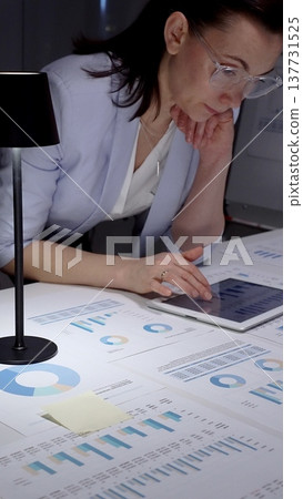 Businesswoman hands analyzing financial data and charts on a digital tablet at an office desk, surrounded by printed reports for marketing strategy and management Businesswoman hands analyzing financial data and charts on a digital tablet at an office desk, surrounded by printed reports for marketing strategy and management 137731525