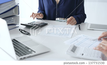 Female financial analysts with male colleague examining documents, using magnifying glass and calculator while examining corporate data at workplace in office. Audit and taxes in business Female financial analysts with male colleague examining documents, using magnifying glass and calculator while examining corporate data at workplace in office. Audit and taxes in business 137731530