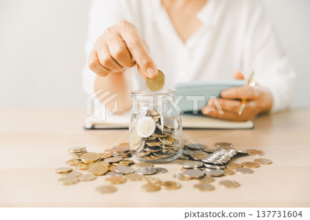Hand dropping coin into glass savings jar on desk with scattered coins, notebook and phone. Budget planning, personal finance, frugal living, investment goals, future security. investment goals 137731604