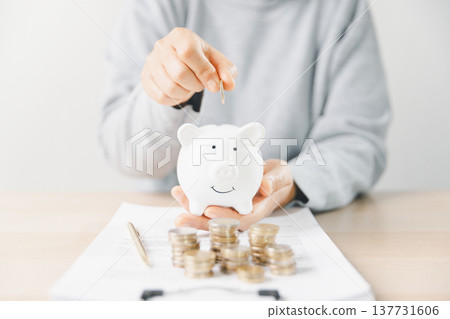 Hand placing a coin into a pink piggy bank beside stacks of money while writing budget notes at home. Personal finance, savings plan, budgeting, investment, long-term future security goals. 137731606