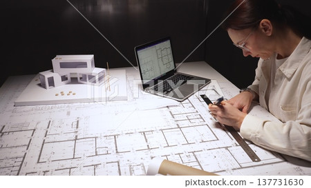 Architect drawing architectural floor plans on a large blueprint paper, working on a complex residential project with a laptop displaying cad designs and a physical house model on the desk Architect drawing architectural floor plans on a large blueprint paper, working on a complex residential project with a laptop displaying cad designs and a physical house model on the desk 137731630
