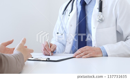 Adult aged doctor man taking notes while listening to a female patient explaining her symptoms during a medical consultation in the clinic office. Medicine and health care concept Adult aged doctor man taking notes while listening to a female patient explaining her symptoms during a medical consultation in the clinic office. Medicine and health care concept 137731635