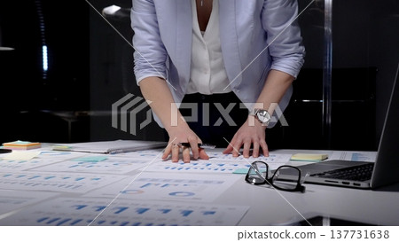 Businesswoman studying financial charts and reports on a desk late at night, analyzing data and strategy for investment, growth and corporate decision making Businesswoman studying financial charts and reports on a desk late at night, analyzing data and strategy for investment, growth and corporate decision making 137731638