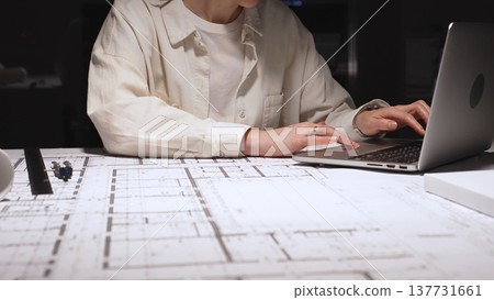 Architect or engineer working late, typing on laptop, reviewing blueprints and technical drawings for a new construction project or building plan in an office Architect or engineer working late, typing on laptop, reviewing blueprints and technical drawings for a new construction project or building plan in an office 137731661