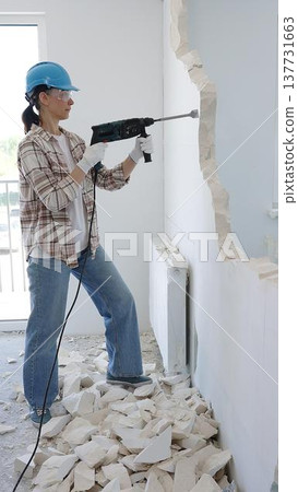 Woman construction worker demolishing a wall with a hammer drill, wearing protective gear, generating rubble pile during renovation project, vertical view 137731663