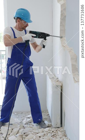 Man construction worker demolishing concrete wall with rotary hammer, wearing protective safety gear, generating dust and debris during home renovation work 137731686