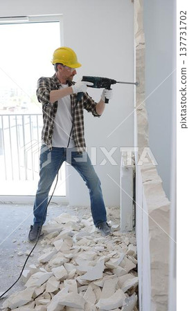 Man construction worker demolishing concrete wall with rotary hammer, wearing safety gear and yellow hardhat, generating rubble and dust during extensive home renovation process 137731702