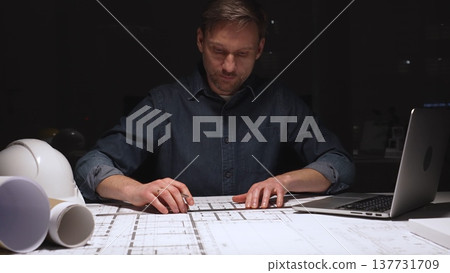 Architect working late into the night, focusing on detailed blueprints and construction plans using a ruler, illuminated by a focused spotlight on the desk 137731709