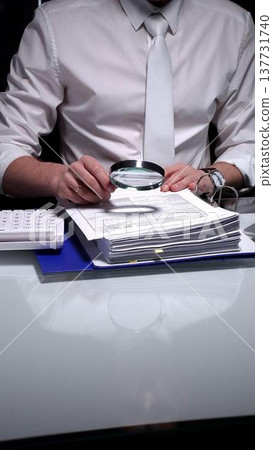 Professional businessman analyzing financial reports and charts with a magnifying glass while working at a desk. Audit and taxes theme in business Professional businessman analyzing financial reports and charts with a magnifying glass while working at a desk. Audit and taxes theme in business 137731740