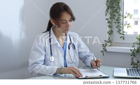 Female doctor wearing a lab coat and stethoscope, diligently writing notes on a clipboard while sitting at a desk in bright medical office with window and decorative plants. Medicine concept. 137731766