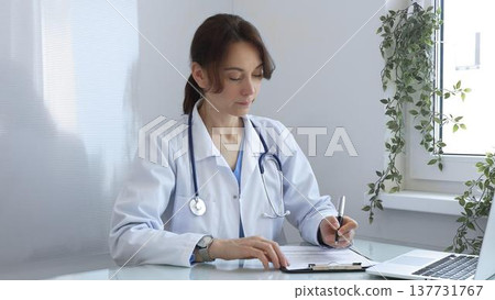 Female doctor wearing a lab coat and stethoscope, diligently writing notes on a clipboard while sitting at a desk in bright medical office with window and decorative plants. Medicine concept. 137731767