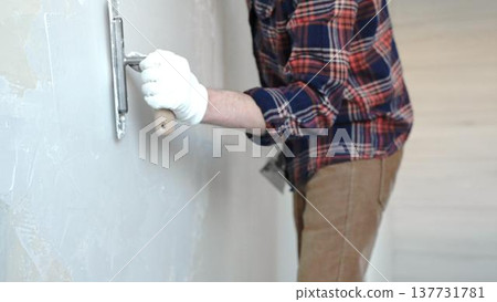 Unknown male construction worker wearing protective gloves smoothing plaster on interior wall during home renovation, applying material with professional trowel technique, close-up Unknown male construction worker wearing protective gloves smoothing plaster on interior wall during home renovation, applying material with professional trowel technique, close-up 137731781