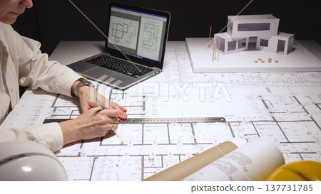 Architect reviewing blueprints and cad on a laptop while sketching a house model and planning construction details at a desk, workspace focused on precision and design, close up Architect reviewing blueprints and cad on a laptop while sketching a house model and planning construction details at a desk, workspace focused on precision and design, close up 137731785