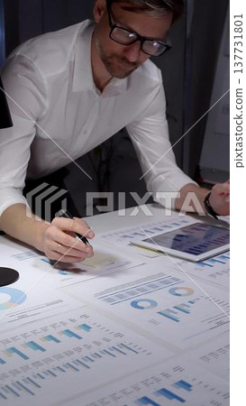 Business professional studies financial reports and market data at a cluttered desk, surrounded by charts, graphs and a laptop, focused on planning, strategy and results Business professional studies financial reports and market data at a cluttered desk, surrounded by charts, graphs and a laptop, focused on planning, strategy and results 137731801
