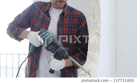 Male construction worker wearing red checkered shirt and protective white gloves is demolishing white wall with rotary hammer drill, closeup view Male construction worker wearing red checkered shirt and protective white gloves is demolishing white wall with rotary hammer drill, closeup view 137731808