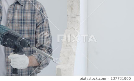 Construction worker demolishing white wall with rotary hammer drill, wearing grey checkered shirt and protective white gloves, generating dust during renovation project, closeup view 137731900