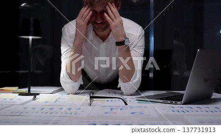 Stressed businessman suffering from overwhelming burnout and work overload, working late into the night, feeling frustrated with deadline pressure in a dark office environment 137731913
